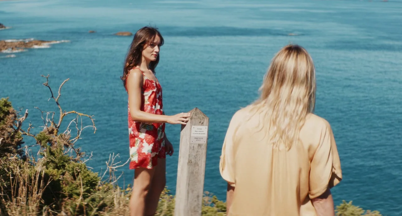 Anaïs in Love (2021) movie still: fullbody — Two women stand on a grassy cliffside overlooking a deep blue ocean body. The woman on the left rests…