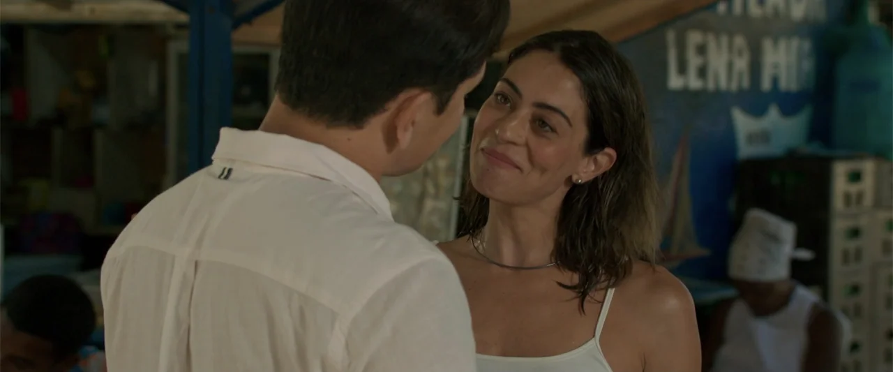 Férias Trocadas (2024) movie still: medium — A dimly lit indoor space with blue wall lettering and stacked storage crates. A man in a white shirt…