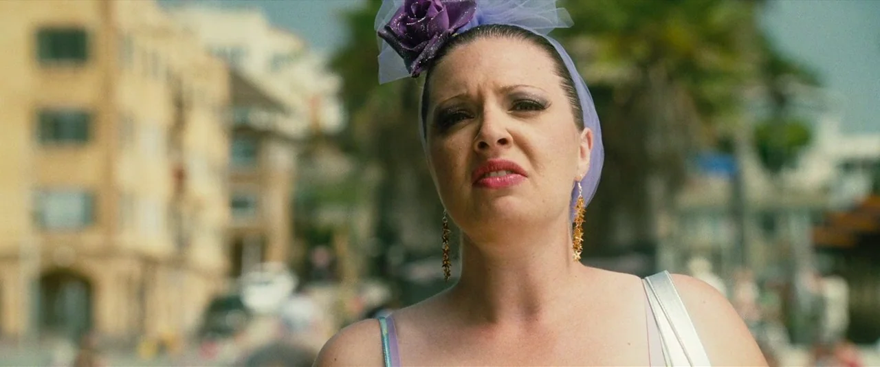 Southland Tales (2006) movie still: closeup — A woman stands outdoors in a public area with blurred buildings and palm trees. She wears a decorative…