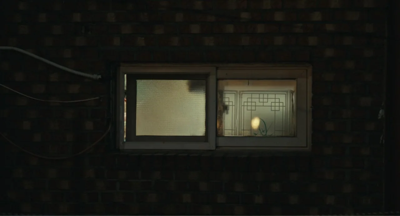 Winter in Sokcho (2025) movie still: medium — A dark brick wall surrounds a dual-pane sliding window with frosted glass inserts. The frame captures…
