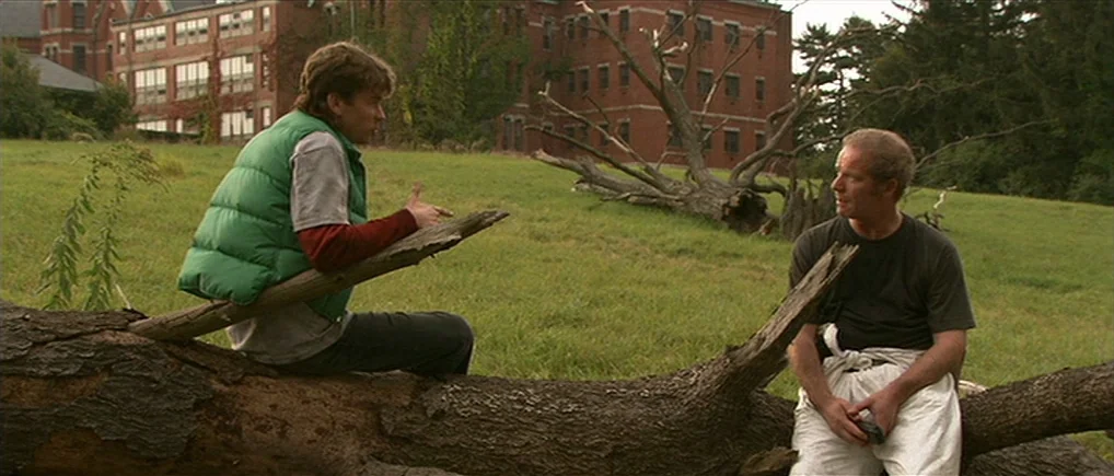 Session 9 (2001) movie still: fullbody — An open grassy field featuring a large brick institutional building in the background. Two men are seated…