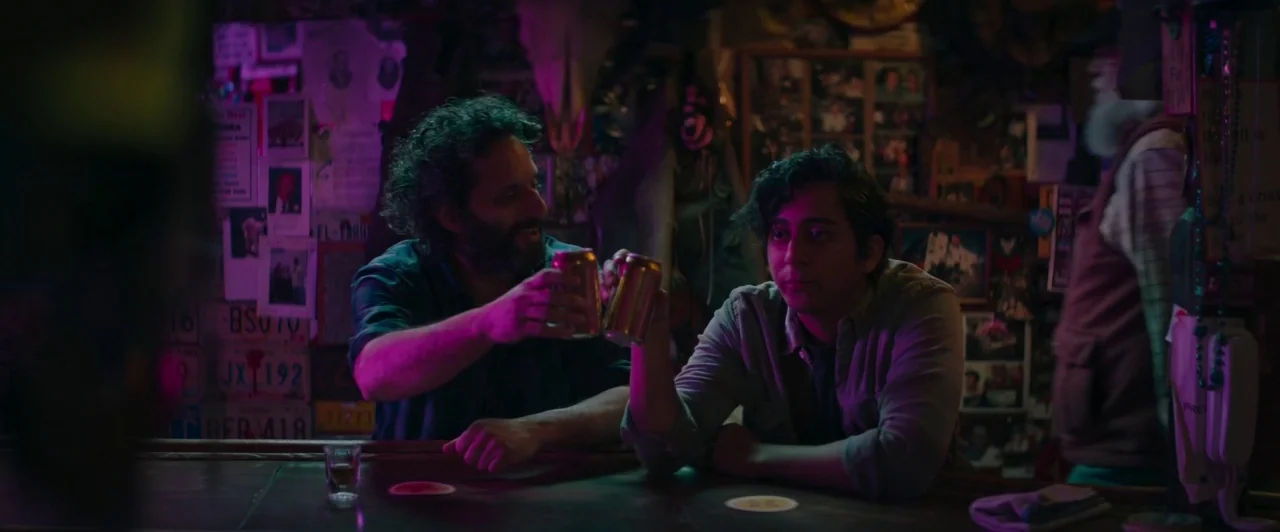 The Long Dumb Road (2018) movie still: medium — A dimly lit, cluttered bar interior features walls covered in vintage license plates and photos. Two…