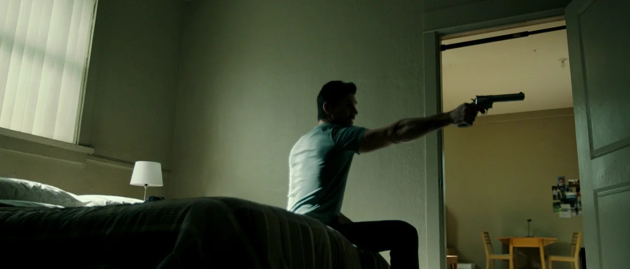 The Purge: Anarchy (2014) movie still: medium — A dimly lit motel room features a bed covered with a textured blanket. A man sits on the edge of the…