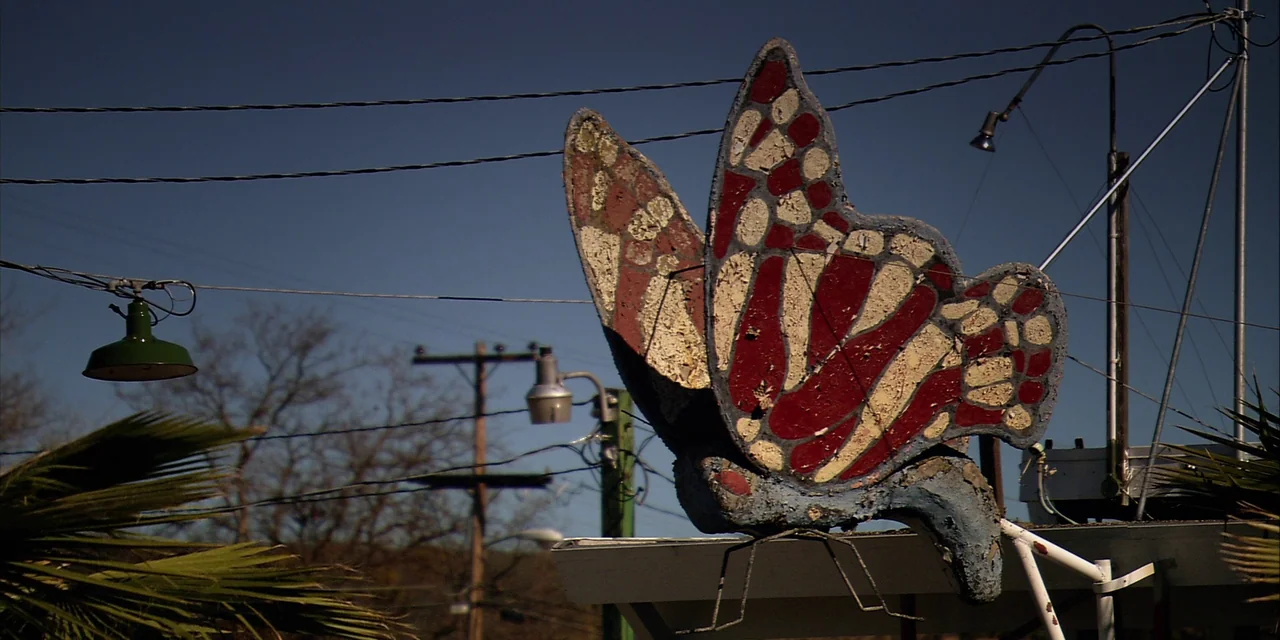 Twixt (2011) movie still: medium — A weathered, painted butterfly sculpture sits mounted on a post against a clear sky. Power lines and utility…