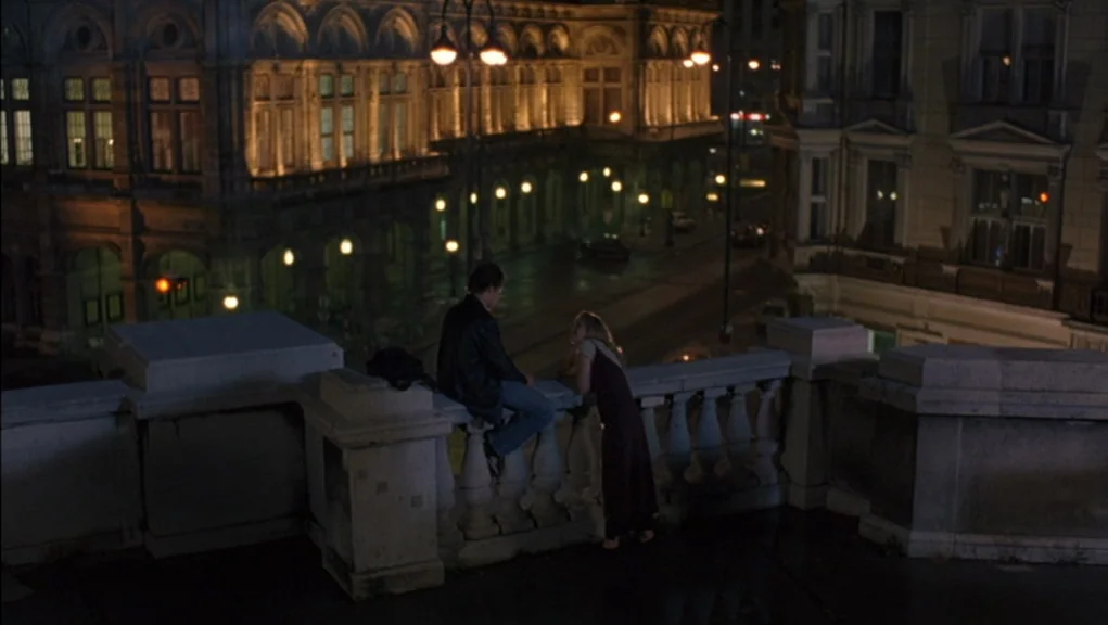 Before Sunrise (1995) movie still: wide — A stone balcony overlooks a quiet, illuminated street flanked by historical ornate buildings at night. A…