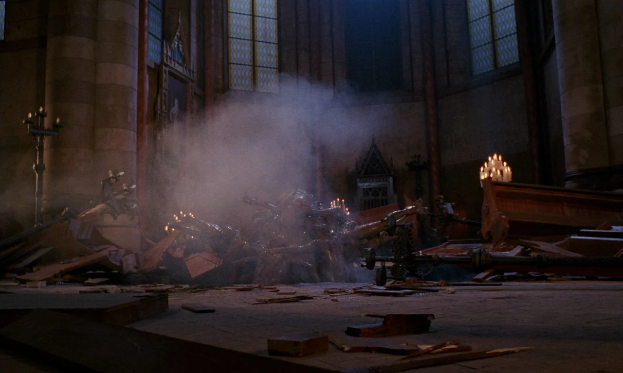 The Church (1989) movie still: wide — A stone cathedral nave shows structural damage with scattered wooden pews and debris. Smoke clouds rise from…