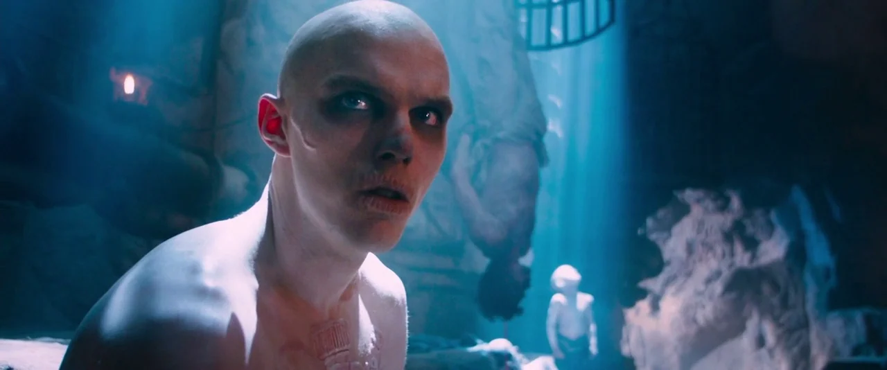 Mad Max: Fury Road (2015) movie still: medium — A dark subterranean space filled with stone structures and hanging figures in deep shadows. A bald…