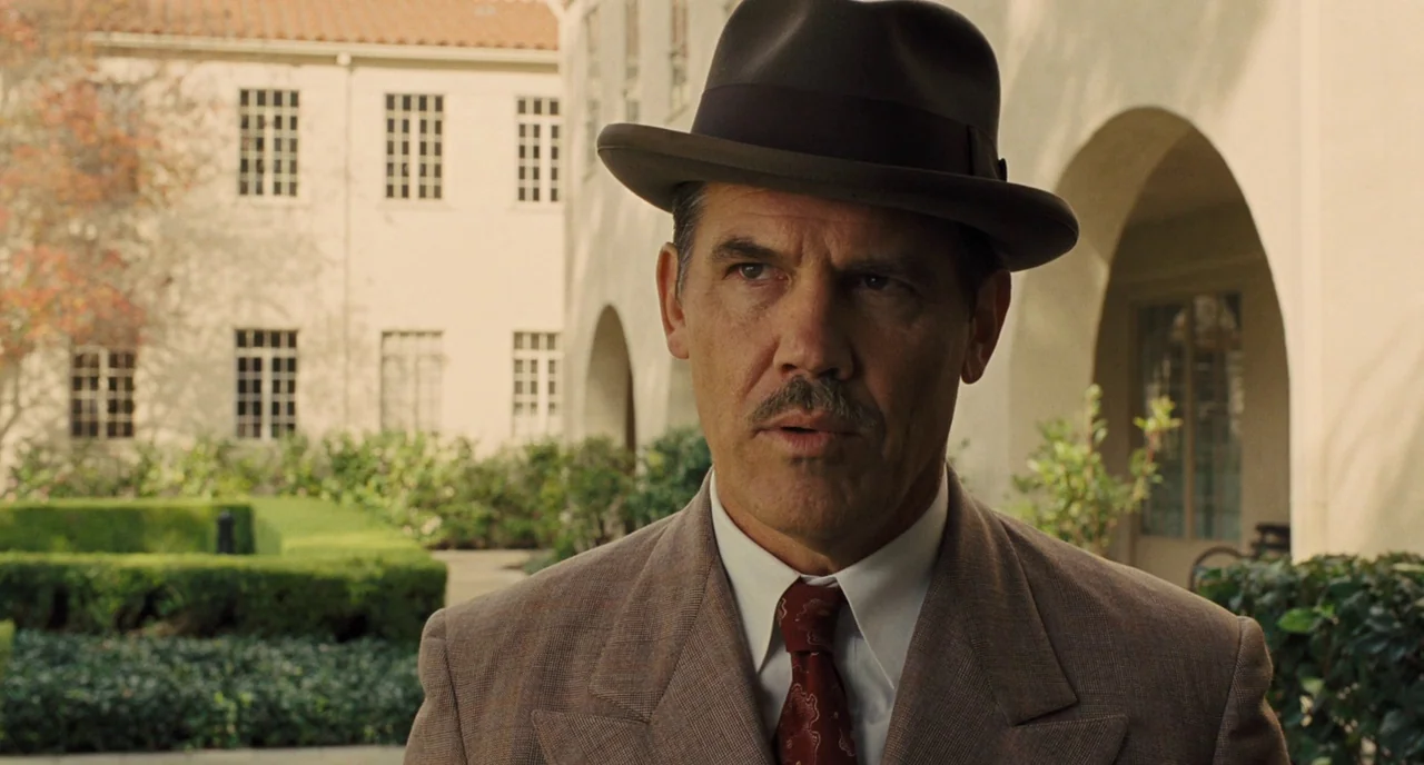 Hail, Caesar! (2016) movie still: medium — A sunny courtyard with Spanish colonial architecture and manicured hedges in the background. A man…