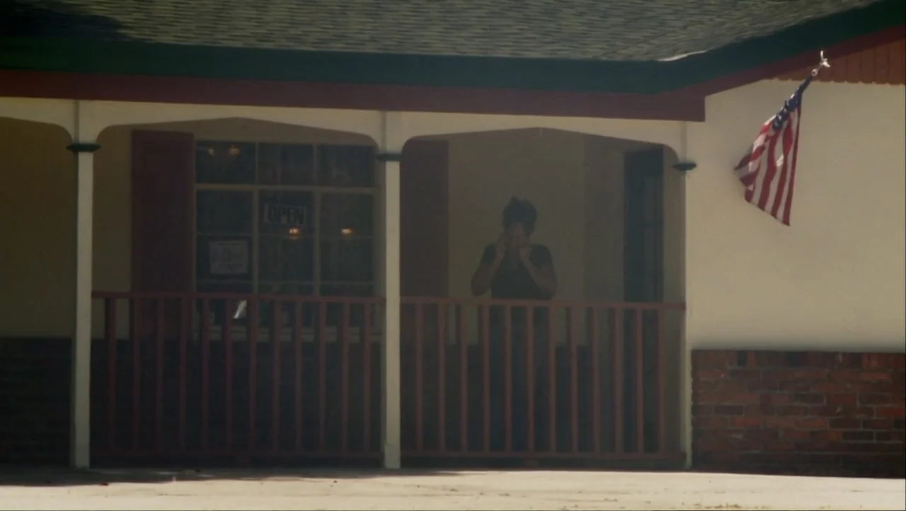 12th & Delaware (2010) movie still: medium — A single-story commercial building exterior features white walls and a reddish-brown porch railing. A…