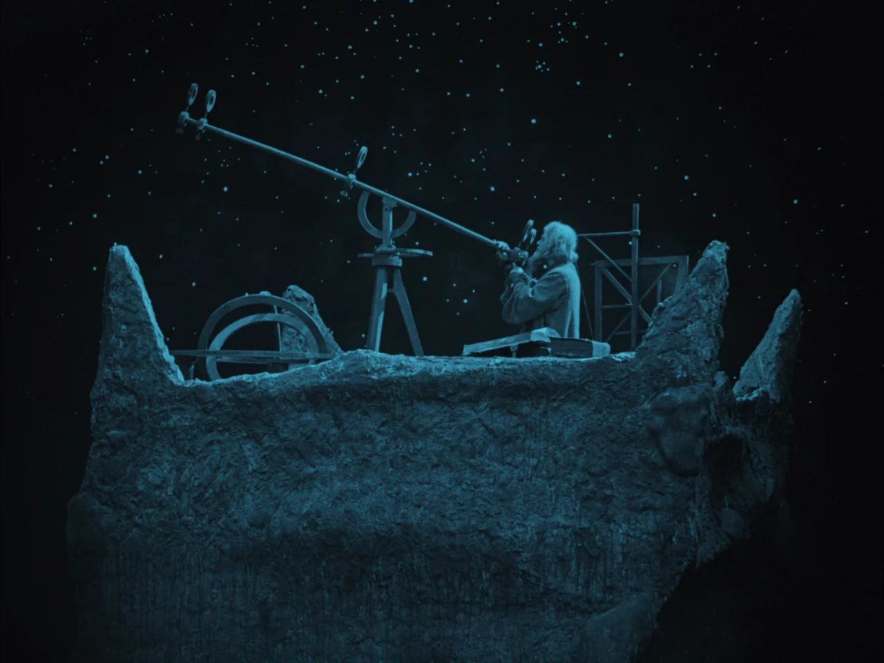The Golem (1920) movie still: wide — A man stands on a craggy, painted rock structure beneath a starry backdrop. He peers through a large vintage…