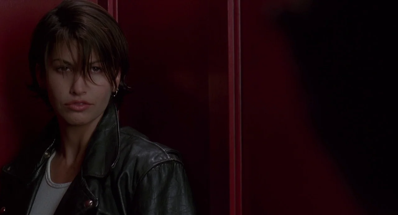 Bound (1996) movie still: medium — A woman stands before a dark red wall with vertical paneling. She gazes slightly off-frame while wearing a…