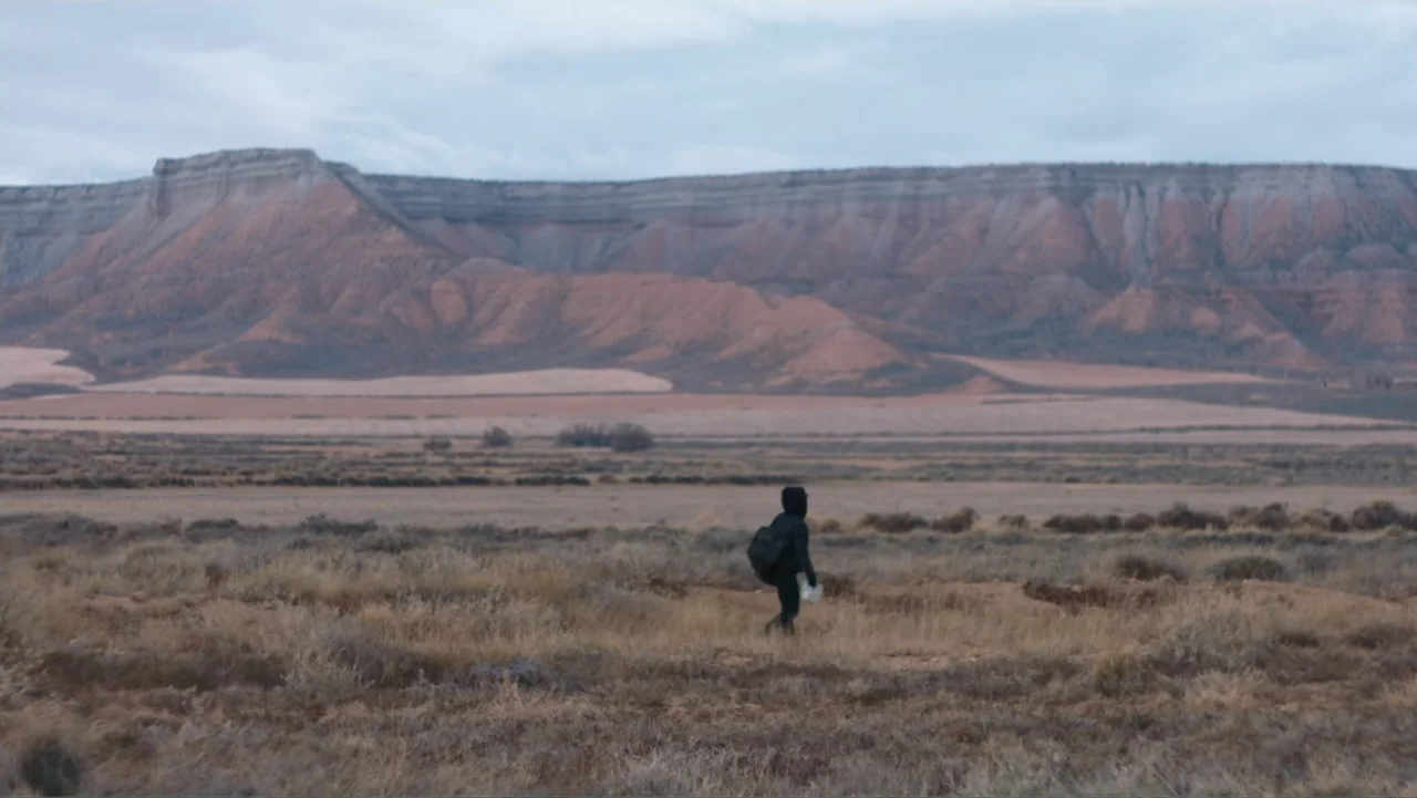 Code of Trust (2019) movie still: wide — An arid, grassy expanse stretches toward a large mesa under a cloudy sky. A person in dark clothing walks…