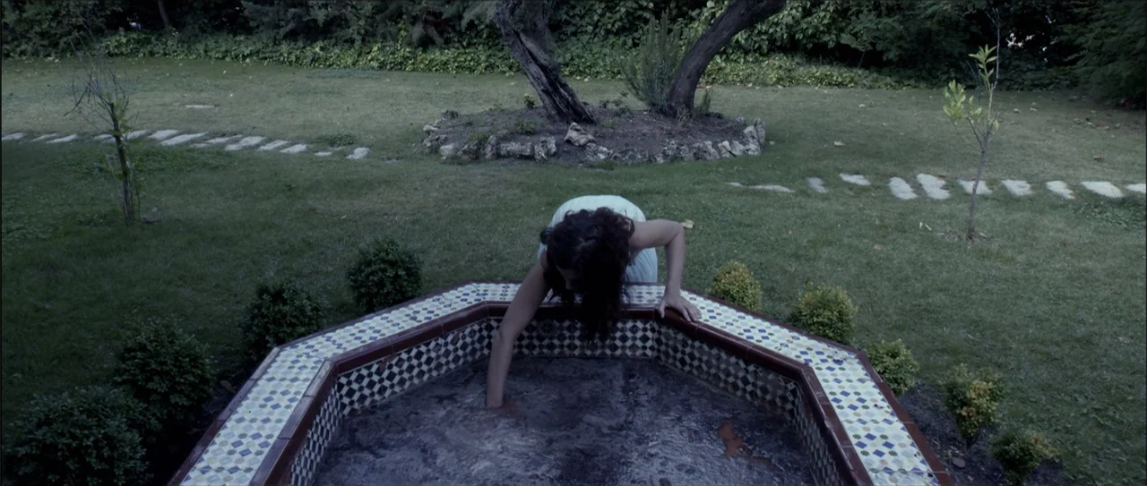 Otro verano (2013) movie still: medium — A dark tiled hexagonal fountain sits in the center of a grassy yard at dusk. A woman in a light dress leans…