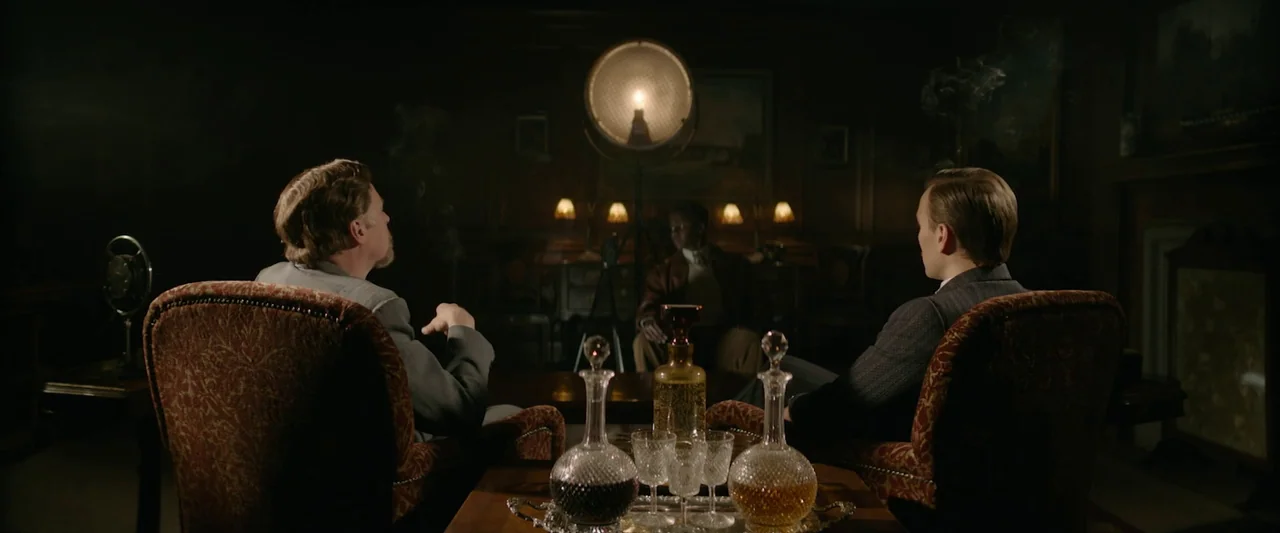Nandor Fodor and the Talking Mongoose (2023) movie still: wide — A dark wood-paneled room features two men seated in the foreground facing a third…