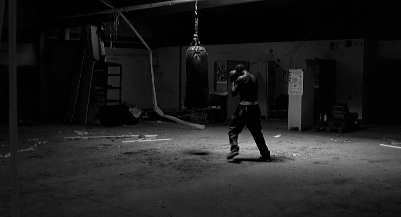La Haine (1995) movie still: wide — An abandoned industrial space has debris scattered across the concrete floor. A shirtless man punches a heavy…