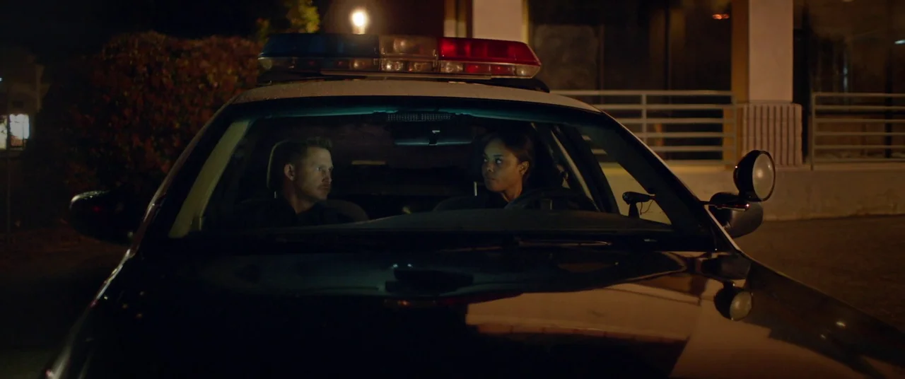 Blindfire (2020) movie still: medium — The interior of a patrol car parked at night with a building in the background. Two police officers sit…