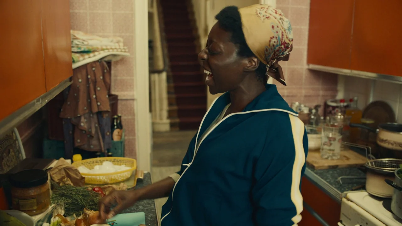Lover’s Rock (2020) movie still: medium — A cramped kitchen with orange cabinets and tiled walls shows a staircase in back. A woman with a headscarf…