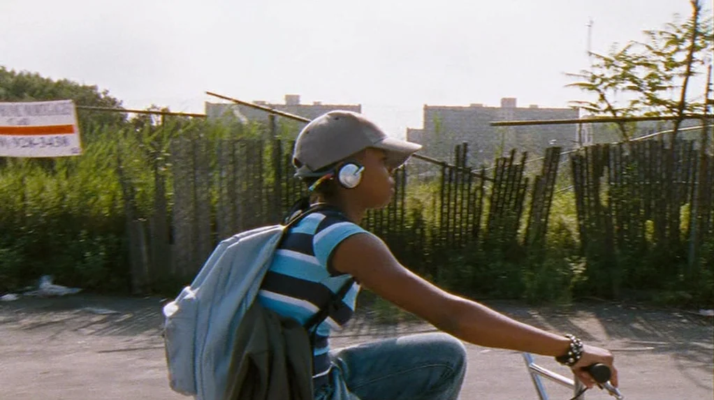 Half Nelson (2006) movie still: medium — An overgrown vacant lot with chain-link fencing borders a paved path in the background. A young person…