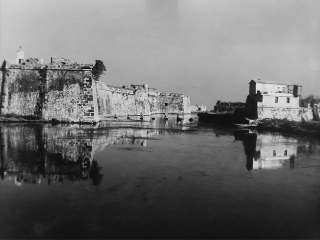 Chained Girls (1965) movie still: establishing — A massive stone fortress wall spans the left side of the frame near water. A smaller white building…