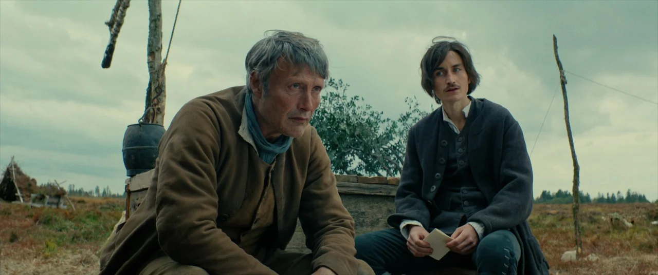 The Promised Land (2023) movie still: medium — Two men sit on a wooden structure amidst a vast, overcast, and desolate moorland. The older man looks…