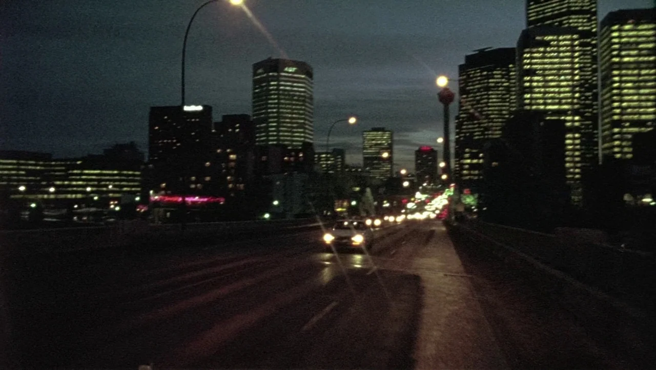 Hard Core Logo movie still: establishing — A dark urban bridge leads toward a skyline filled with illuminated office towers. A lone car approaches…