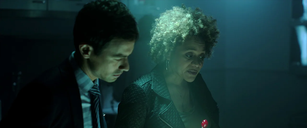 Malignant (2021) movie still: medium — A dimly lit room with cool green light reflects off metallic and dark surfaces. A man and a woman stand…