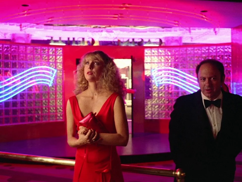 One From The Heart (1982) movie still: medium — A casino interior features glass block walls illuminated by wavy blue neon lights. A woman in a red…