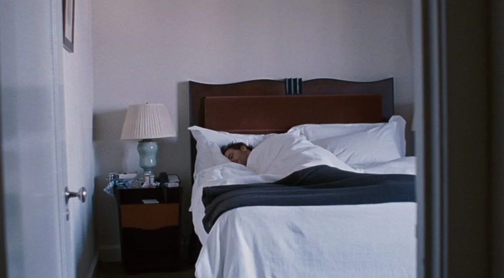 Somewhere (2010) movie still: wide — A hotel room features a dark wood bed frame and a bedside table. A man sleeps under white sheets while the room…