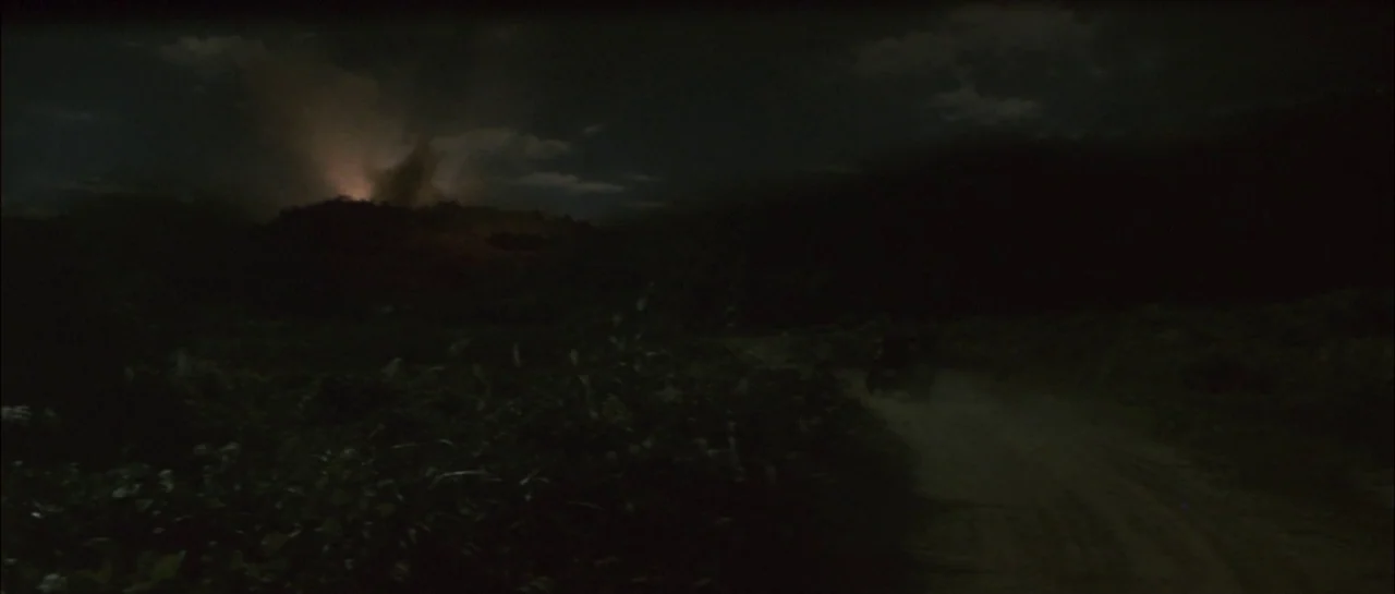 Godzilla Vs Megalon (1973) movie still: wide — A dark rural landscape at night features a dirt path winding through tall grasses. In the distance, a…