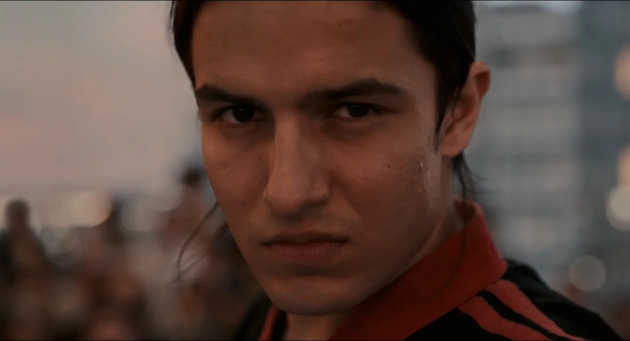 Karate Kid: Legends (2025) movie still: closeup — An outdoor area with a blurred city skyline visible in the background. A young man looks forward…