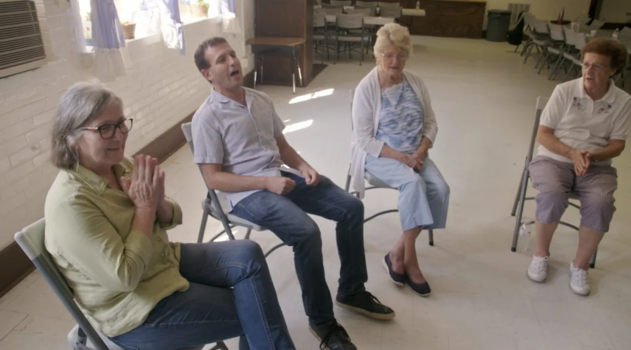 The Divided Soul of America (2019) movie still: fullbody — A bright room with white walls, folding chairs, and tables in the background. Four…