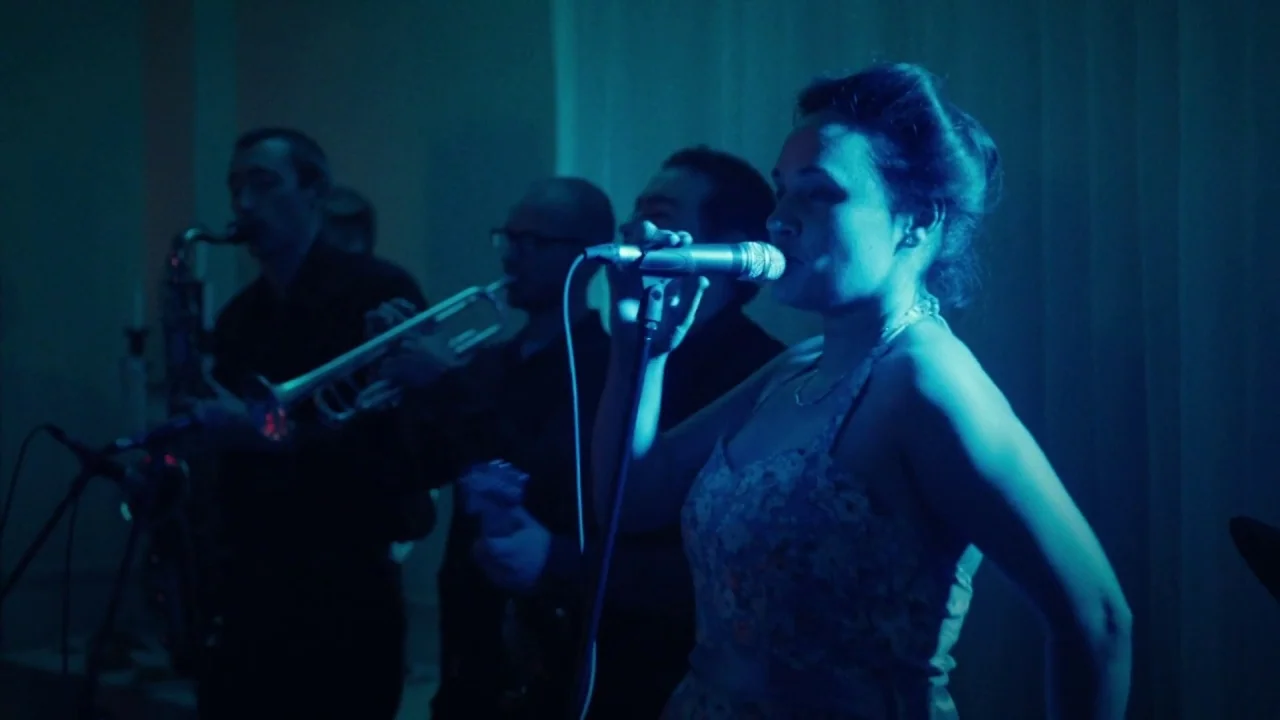 A Date for Mad Mary (2016) movie still: medium — A dimly lit indoor space featuring a musical ensemble performing on a small stage. A woman stands…