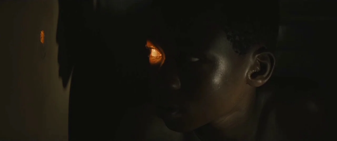 Beasts of No Nation (2015) movie still: closeup — A young boy remains in complete darkness inside a confined, unlit interior space. He looks through…