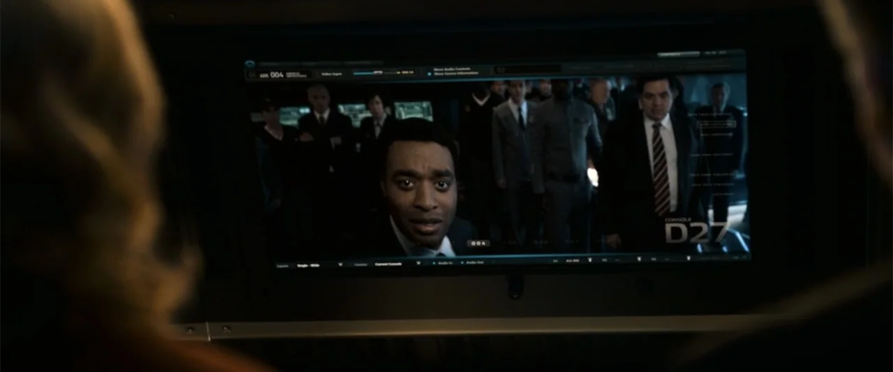 2012 (2009) movie still: medium — A high-tech darkened command center features a large monitor displaying a live video feed. A man in a suit speaks…