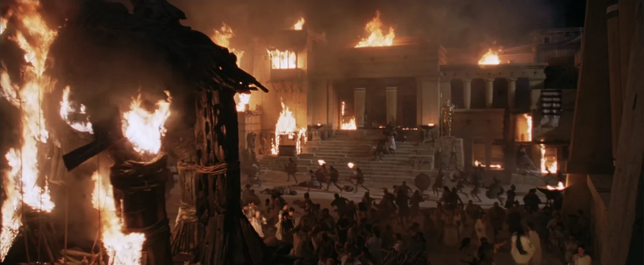 Troy (2004) movie still: wide — A stone courtyard with classical architecture is engulfed in widespread structural fires. A panicked crowd of people…