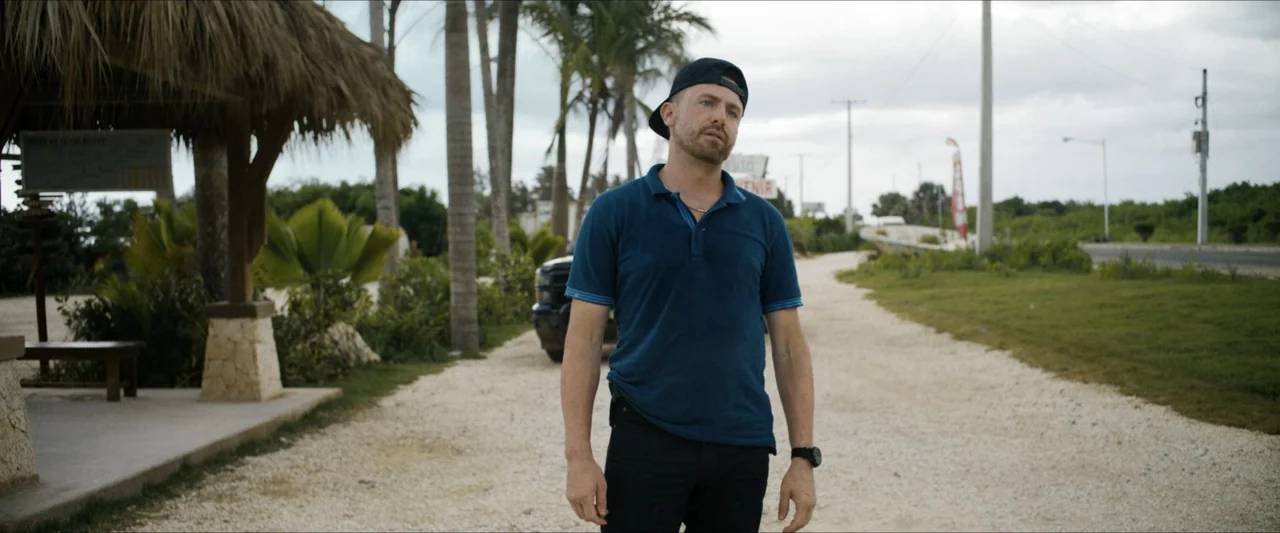 Road House (2024) movie still: fullbody — A gravel roadside path leads through tropical trees toward a distant paved road. A man in a blue polo…