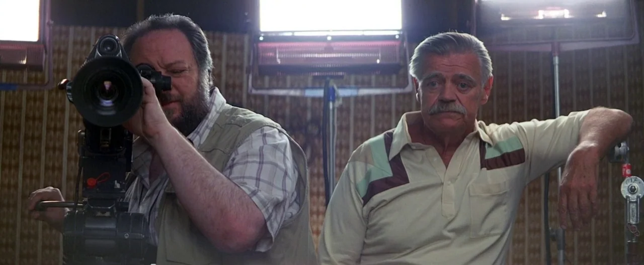 Boogie Nights movie still: medium — Two men stand before large lighting rigs mounted on metal stands against a textured wall. The cameraman peers…