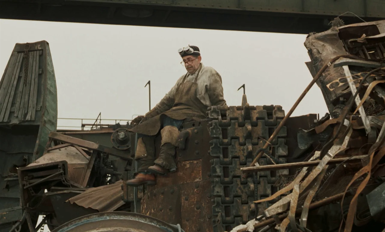 Larks On A String (1969) movie still: fullbody — A man sits atop a large pile of rusted industrial debris and scrap metal. An elevated metal bridge…