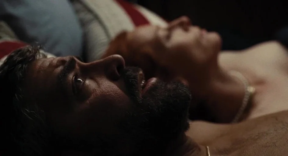 Burn After Reading (2008) movie still: closeup — A dark, blurred sleeping area with pillows and bedding partially visible in the background. A man…