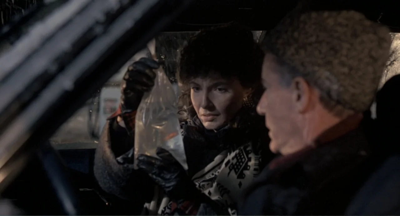 Dead of Winter (2025) movie still: medium — Two individuals sit inside a dark vehicle at night during a rainstorm. The woman displays a clear…