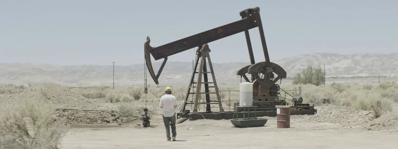 Entertainment (2015) movie still: wide — A dry desert landscape features a large, weathered industrial oil pump jack. A lone man wearing a white…