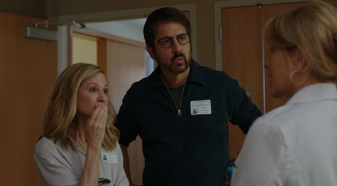 The Big Sick (2017) movie still: medium — A hospital hallway features plain wood doors and beige walls. Two people stand listening to a professional…