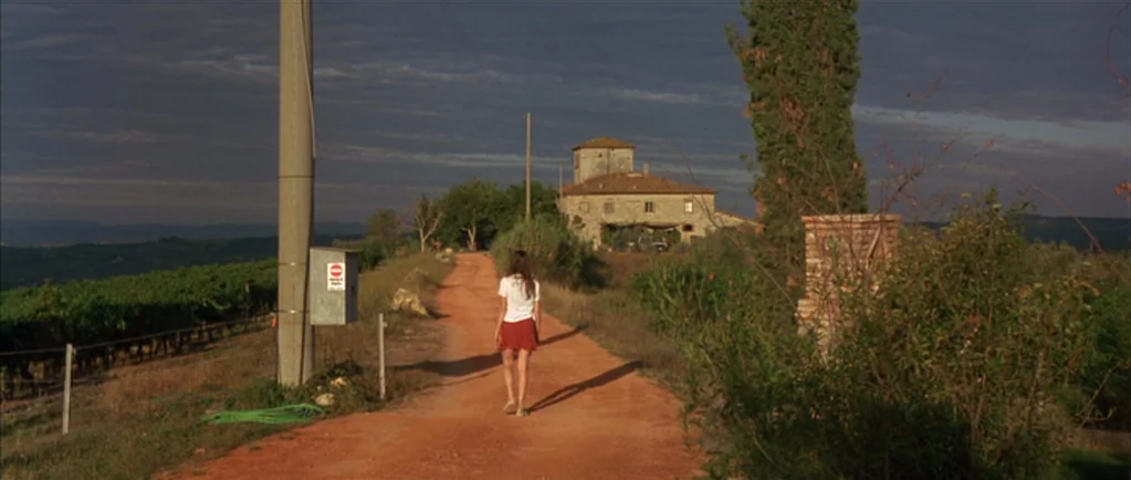 Stealing Beauty (1996) movie still: fullbody — A rural dirt road winds through vineyards toward an old stone house. A woman walks alone down the…