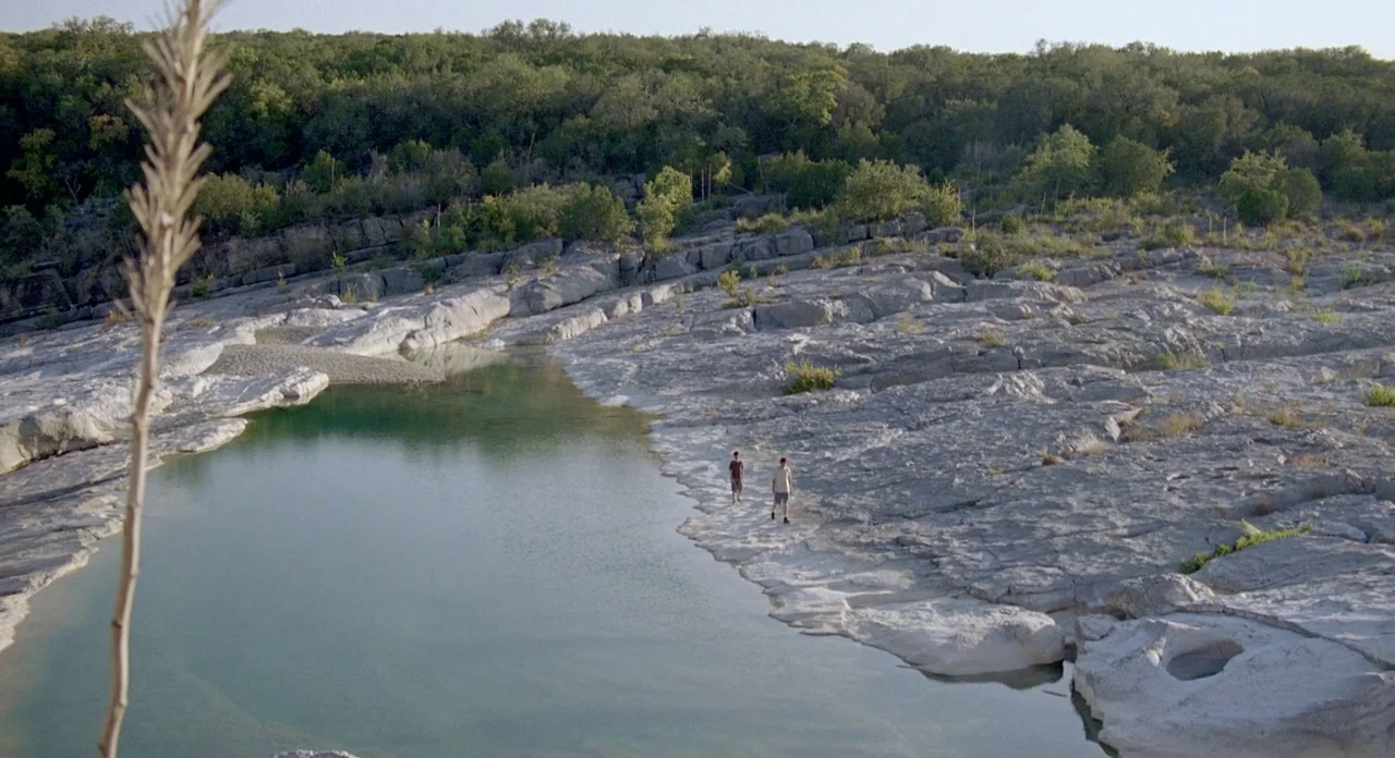 Boyhood (2014) movie still: wide — A vast limestone creek bed features a shallow green pool surrounded by rugged rock. Two teenage boys walk along…