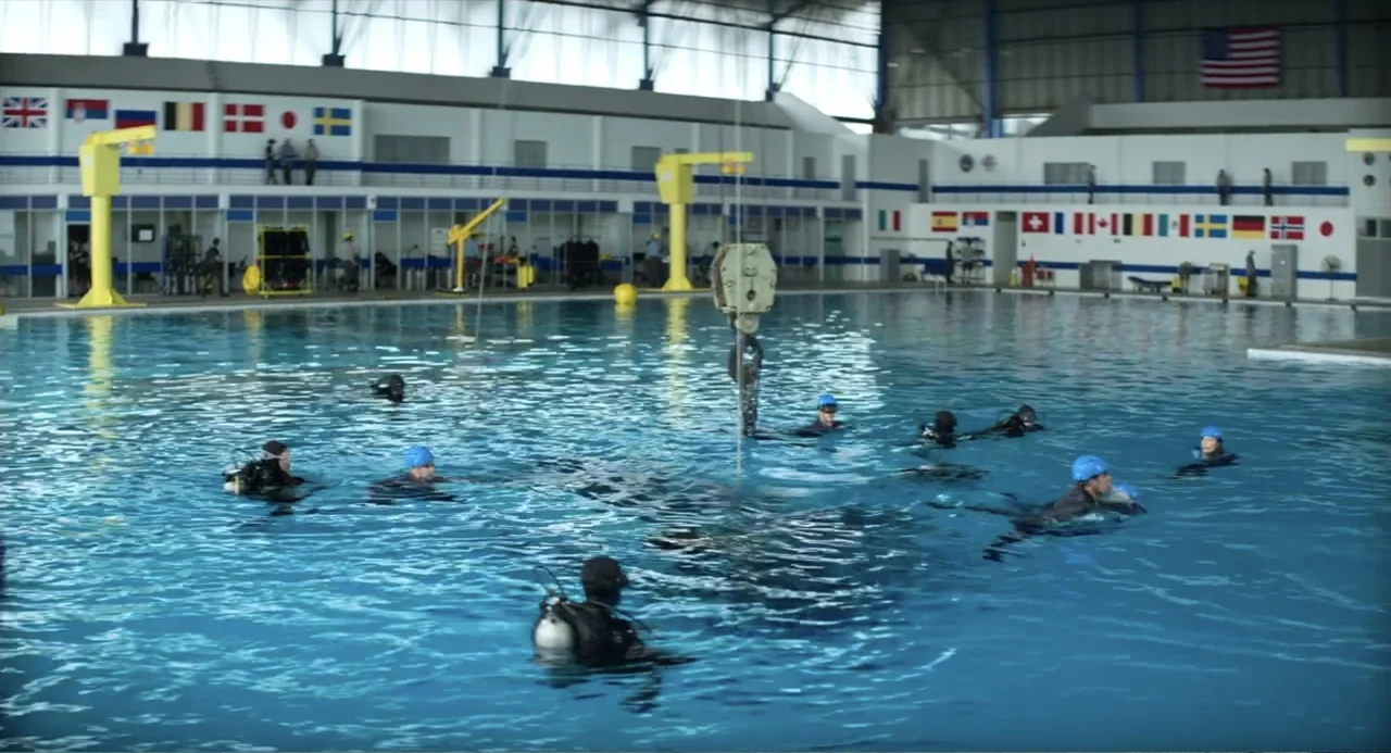 A Million Miles Away (2023) movie still: wide — A large indoor training facility features a blue water tank surrounded by high ceilings. Multiple…