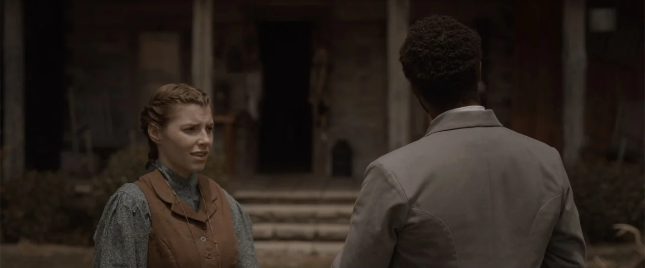 Ghosts of the Ozarks (2022) movie still: medium — A woman speaks to a man standing on the wooden porch of a house. The man has his back to the…