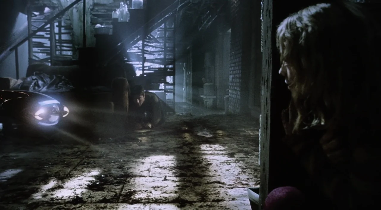 War of the Worlds (2005) movie still: wide — A dark, dilapidated basement with debris scattered across the floor and wooden stairs. A man crawls…