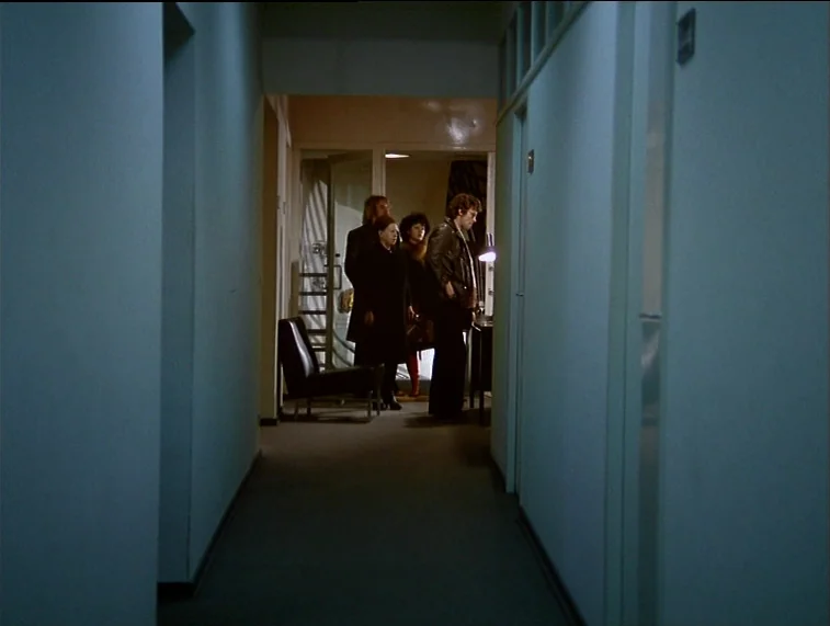 Mother Kusters Goes To Heaven (1975) movie still: wide — An empty narrow hallway with pale walls leads to a brightly lit room. Four people stand…