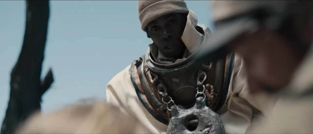 Fear Below (2025) movie still: medium — A sunny beach with a dark, weathered wooden post in the foreground. A man wearing an antique diving suit…