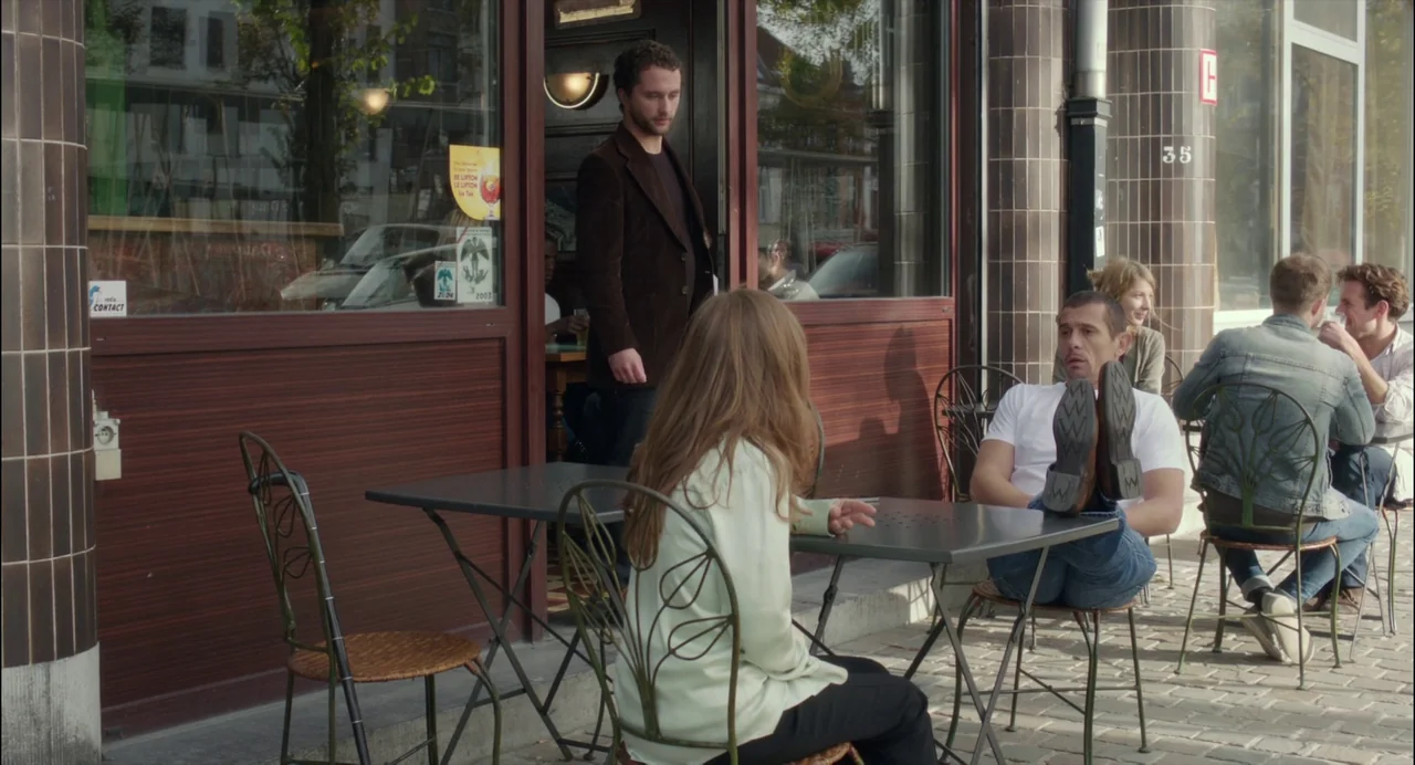 Abuse of Weakness (2013) movie still: fullbody — An outdoor cafe area with metal tables and chairs set against a brown facade. A man relaxes with…